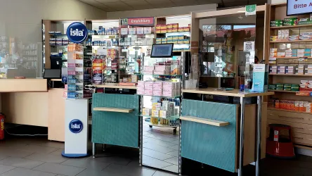 Eine Apotheke mit einem Empfangstresen und Regalen voller Medikamente. An der Wand sind verschiedene Produkte zur Gesundheit und Pflege ausgestellt.