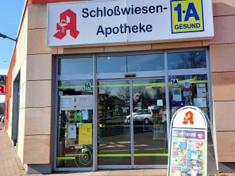 Eine Apotheke mit dem Namen "Schloßwiesen-Apotheke" ist zu sehen. Der Eingang ist gut sichtbar und es gibt ein Werbeschild im Vordergrund.