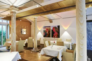 Ein elegantes Restaurant mit weißen Tischtüchern und Holzdetails. An den Wänden hängen bunte Bilder, die dem Raum eine künstlerische Note verleihen.