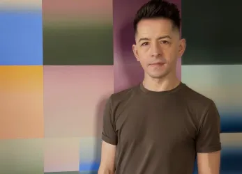Ein Mann steht vor einer farbigen Wand mit geometrischen Mustern. Er trägt ein schlichtes, graues T-Shirt und hat einen modernen Haarschnitt.