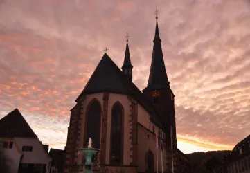 Eine Kirche mit zwei hohen Türmen steht vor einem farbenfrohen Sonnenuntergang. Der Himmel ist mit Wolken in warmen Farbtönen gefüllt.