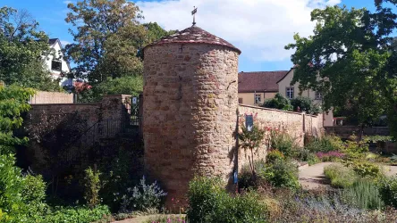 Ein historischer Steinturm mit einem runden Dach in einer grünen Umgebung. Im Hintergrund sind Bäume und ein Gebäude sichtbar.