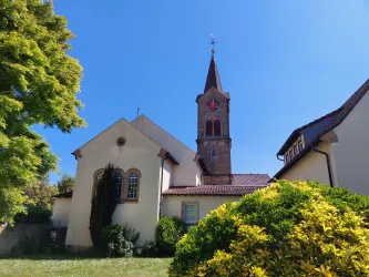 Eine Kirche mit einem hohen Kirchturm steht im Vordergrund. Der Himmel ist klar und blau, umgeben von grünem Gras und Bäumen.