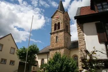 Eine alte Kirche mit einem markanten Turm und einer Uhr. Im Vordergrund sind grüne Bäume und traditionelle Gebäude zu sehen.