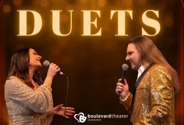 Zwei Sänger stehen sich gegenüber und singen in ein Mikrofon. Der Hintergrund ist festlich mit goldenen Lichtern.