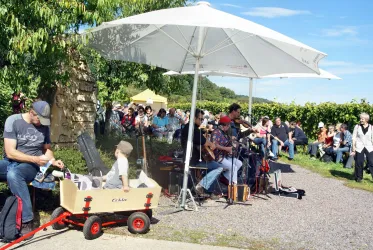 Eine Band spielt Musik unter einem Sonnenschirm, umgeben von Publikum. Im Vordergrund ist eine Person mit einem Wagen zu sehen, der ein Kind und Musikinstrumente hält.