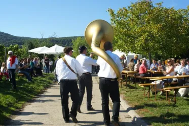 Eine Festveranstaltung im Freien mit Menschen, die an Tischen sitzen. Musikanten spielen eine Tuba und tragen traditionelle Kleidung.