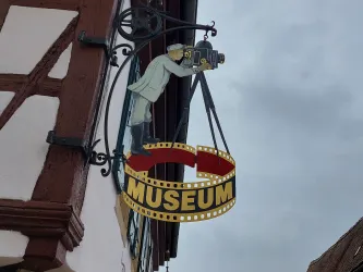 Ein dekoratives Schild mit einem Fotografen, der eine Kamera hält. Das Schild zeigt den Schriftzug "MUSEUM" und hängt an einem Gebäude.