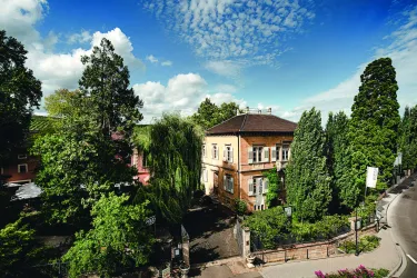 Ein historisches Gebäude umgeben von üppigem Grün und Bäumen. Der Himmel ist blau mit einigen Wolken.