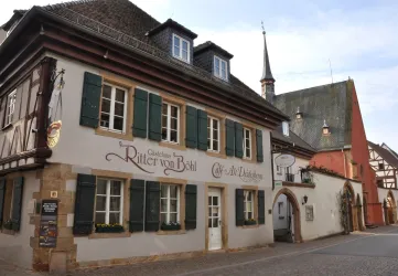 Ein charmantes historisches Gebäude mit grünem Fensterladen und auffälliger Beschilderung. Im Hintergrund sind weitere traditionelle Gebäude und ein Kirchturm sichtbar.