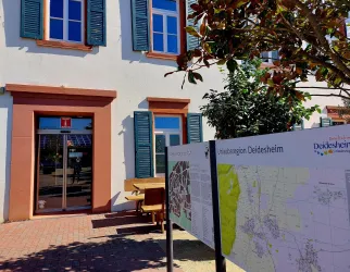 Ein Bürogebäude mit einem großen Fenster und grünen Fensterläden. Vor dem Gebäude steht eine Informationskarte über die Region Deidesheim.