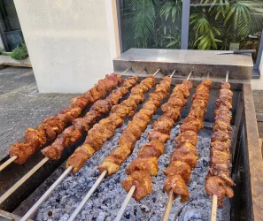 Eine Reihe von Grillspießen mit mariniertem Fleisch auf einem Grill. Im Hintergrund sind Pflanzen und ein Gebäude sichtbar.