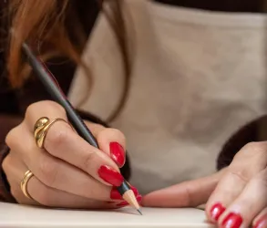 Eine Hand mit rotem Nagellack hält einen Bleistift und zeichnet auf Papier. Im Hintergrund ist ein graues Hemd oder eine Schürze zu sehen.
