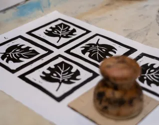 Eine Druckgrafik mit vier schwarzen Blattmustern auf weißem Papier. Daneben liegt ein Holzstempel, der zum Bedrucken verwendet wurde.