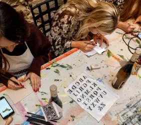 Eine Gruppe von Frauen sitzt an einem Tisch und zeichnet. Auf dem Tisch liegen Stifte, Papier und eine Flasche Wein.