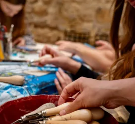 Eine Gruppe von Menschen arbeitet an einem kreativen Projekt. Im Vordergrund sind verschiedene Werkzeuge und Hände zu sehen, die aktiv beim Arbeiten sind.