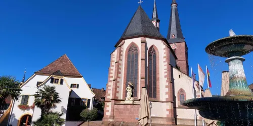 Eine beeindruckende Kirche mit hohen Türmen vor klarem blauen Himmel. Im Vordergrund befindet sich ein schöner Brunnen und palmenbesäumte Gebäude.