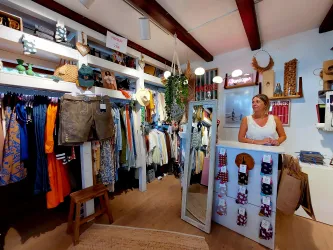 Ein einladender Bekleidungsraum mit bunten Kleidungsstücken und Accessoires. Eine freundliche Verkäuferin steht an der Kasse und blickt in die Kamera.