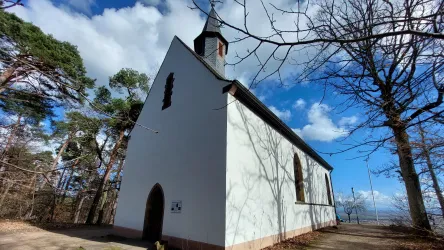 Eine kleine weiße Kirche steht umgeben von Bäumen unter einem blauen Himmel. Die Architektur zeigt einen schlichten und charmanten Stil.
