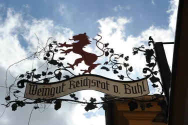 Ein Schild des Weinguts Reichsrat von Buhl, dekoriert mit einem roten Löwen und Weinranken. Der Himmel ist teilweise bewölkt.