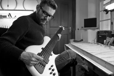 Ein Mann sitzt und spielt einen weißen Bass. Im Hintergrund sind Werkzeuge und ein Bildschirm sichtbar.