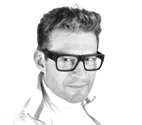 Ein Mann mit kurzen, lockigen Haaren und einer sportlichen Jacke. Er trägt große, auffällige Brillen und schaut direkt in die Kamera.