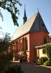 Eine kleine Kirche mit einem spitzen Turm und bunten Fenstern. Die Fassade ist in warmen Farben gehalten und von Bäumen umgeben.