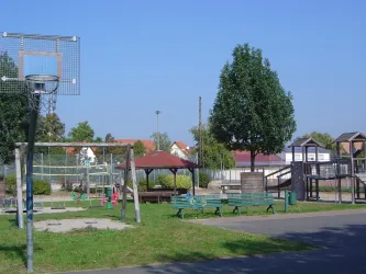 Ein Spielplatz mit einem Basketballkorb, Schaukeln und einem Spielhaus. Im Hintergrund sind Bäume und eine klare blaue Himmel zu sehen.