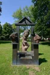 Eine Skulptur in einem Park zeigt eine Figur in der Mitte, flankiert von zwei Tierköpfen. Der Hintergrund besteht aus grünen Bäumen und einem klaren Himmel.