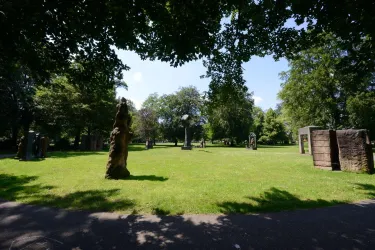 Ein sonniger Park mit grünem Rasen und mehreren Skulpturen. Umgeben von Bäumen bietet der Platz eine entspannte Atmosphäre.