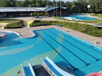 Ein großes Schwimmbad mit mehreren Wasserflächen und Rutschen. Die Atmosphäre ist hell und einladend, umgeben von Bäumen.