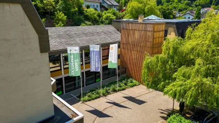 Ein moderner Gebäudekomplex mit großen Fenstern und einem grünen Innenhof. Sichtbare Fahnen und üppige Bäume verleihen der Umgebung einen einladenden Charakter. Hier ist die Biosphärenakademie von Lambrecht (Pfalz) zu sehen.
