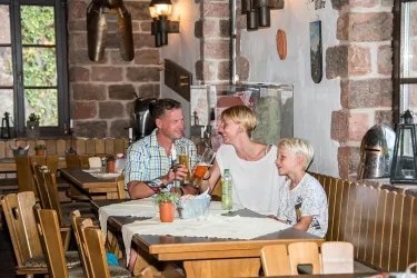 Eine gemütliche Gaststätte mit Holzstühlen und einem freundlichen Ambiente. Eine Familie genießt Getränke und das Zusammensein am Tisch. Hier ist der Gastraum der Burgschänke Burg Spangenberg zu sehen.