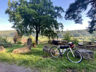 Ein Gravelbike steht an einem Sandstein angelehnt, vor einem schönen Ausblick auf eine grüne Landschaft. Im Hintergrund sind Bäume und Sitzgelegenheiten zu sehen.
