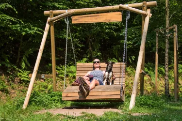 Eine Person entspannt auf einer Wellenliege im Freien. Neben ihr sitzt ein Hund in der Sonne.