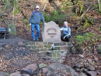 Zwei Personen posieren neben dem Hengstentalbrunnen aus Sandstein im Wald. Der Brunnen trägt die Aufschrift „HENGSTENTAL BRUNNEN 2020“.