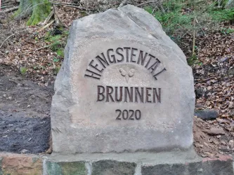 Ein großer, gravierter Stein mit der Aufschrift "Hengstental Brunnen 2020". Der Stein steht in einer natürlichen Umgebung mit Laub und Bäumen.