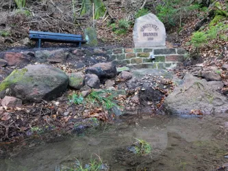 Eine Brunnenanlage mit einem Sandstein und der Inschrift Hengstentalbrunnen 2020, umgeben von Natur. Im Vordergrund gibt es Wasser und einige große Steine.