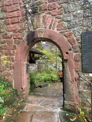 Ein historischer Torbogen aus rötlichem Stein, der in das Innere der Burg Spangenberg führt. Im Hintergrund befindet sich eine ruhige Szenerie aus Natur und Sitzgelegenheiten.