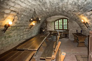 Ein rustikaler Raum der Burgschänke Burg Spangenberg mit steinerner Decke und Holztischen, warm erleuchtet von Wandleuchten und Kronleuchter – gemütlich und einladend.
