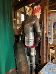 Ein historischer Ritter mit einer Rüstung steht neben einer Wand. Er hält ein Schild und hat eine Feder auf seinem Helm. Die abgebildete Ritterrüstung befindet sich in der Burgschänke Burg Spangenberg.