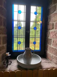 Eine stilvolle Fensterbank mit bunten Glasfenstern und einem Krug auf einer Schale. Im Hintergrund ist die grüne Landschaft sichtbar. Das bunte Glasfenster befindet sich in der Burgschänke Burg Spangenberg.