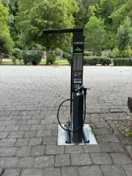 Eine Fahrradreparaturstation mit Luftpumpe und Werkzeugen steht auf einem gepflasterten Weg. Im Hintergrund sind grüne Bäume und eine parkähnliche Umgebung zu sehen.