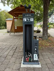 Eine Fahrradreparaturstation mit verschiedenen Werkzeugen und einer Luftpumpe. Sie befindet sich in einem öffentlichen Bereich mit Bäumen im Hintergrund.
