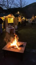 Ein gemütliches Feuer brennt in einer Feuerstelle. Im Hintergrund sind festlich dekorierte Stände und Bäume zu sehen.