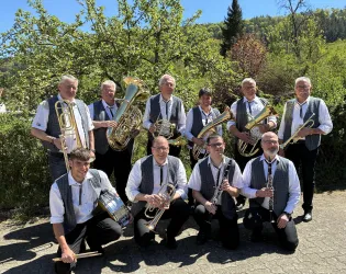 Eine Blaskapelle bestehend aus zehn Musikern zeigt sich lächelnd im Freien. Sie spielen verschiedene Blechblasinstrumente vor einer grünen Landschaft.
Zu sehen ist auf dem Foto der Musikverein Elmstein e.V.