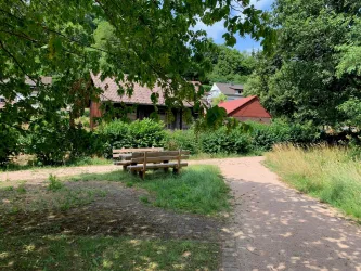 Ein ruhiger Weg in der Natur mit Bänken und üppigem Grün. Im Hintergrund sind einige idyllische Gebäude zu sehen.