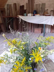 Ein schöner Strauß mit bunten Wildblumen steht im Vordergrund. Im Hintergrund sind Tisch und Geschirr in einer rustikalen Umgebung zu sehen.
