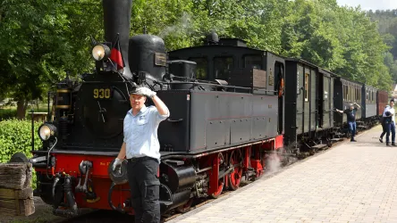 Die dampfbetriebene Lokomotive des Kuckucksbähnels steht am Kuckucksbahnhof in Elmstein. Ein Arbeiter begrüßt die Passagiere neben den Waggons in einer grünen Umgebung.