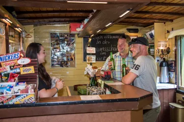 Die einladende Theke in der Museumsschänke des Kuckucksbähnels, mit einem freundlichen Team. Zwei Männer unterhalten sich am Tresen, während sie von einer Frau bedient werden.